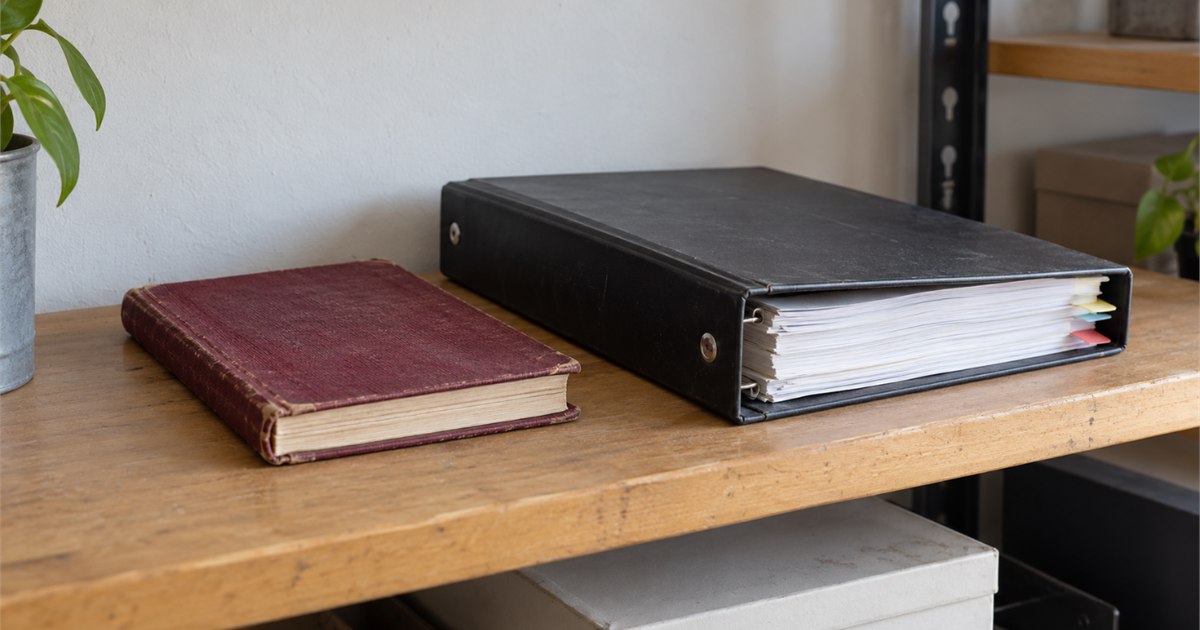 Un carnet rouge usé à côté d’un classeur noir à anneaux sur une tablette en bois